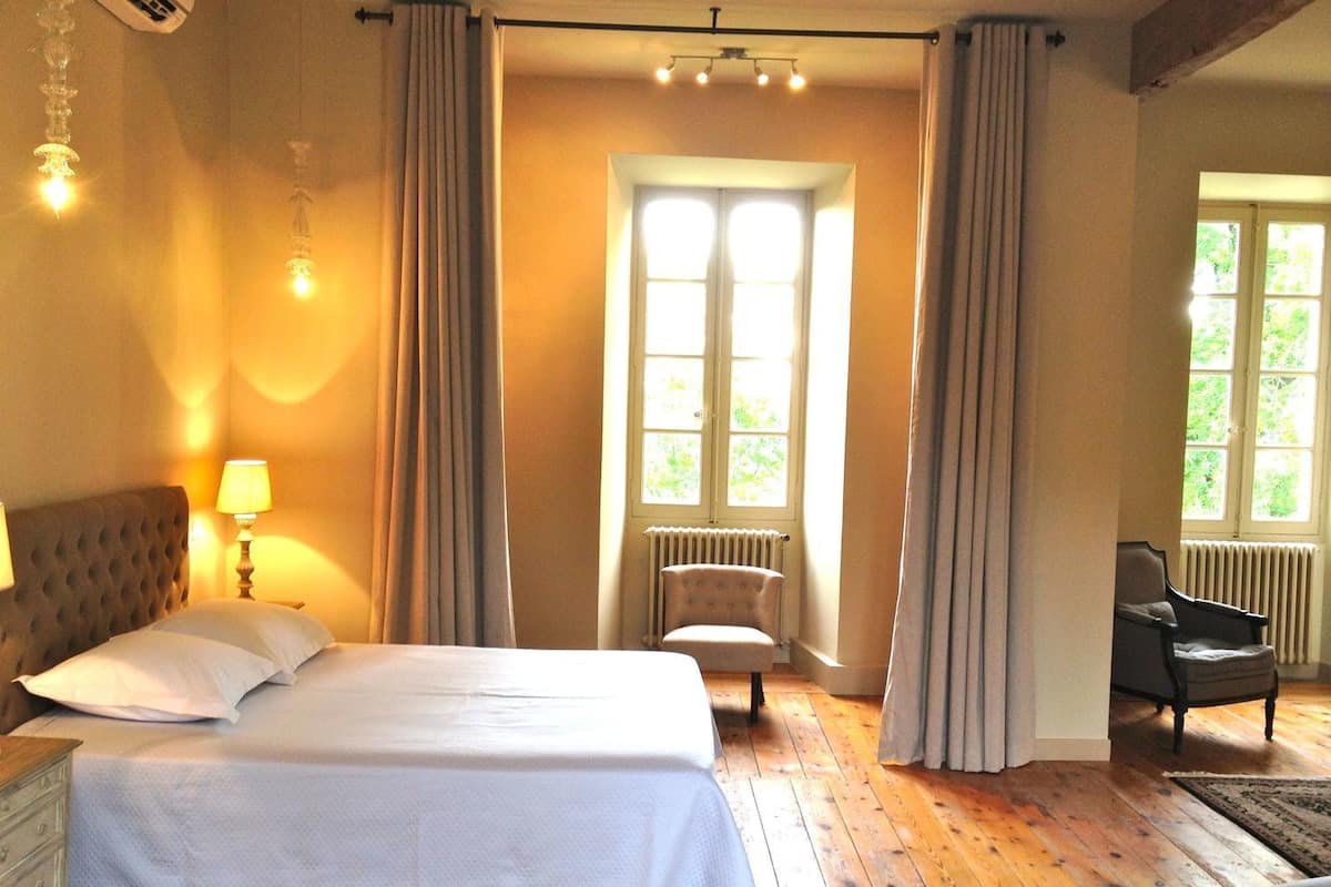 A bedroom with a large bed, a chair, a desk, and a window with curtains.