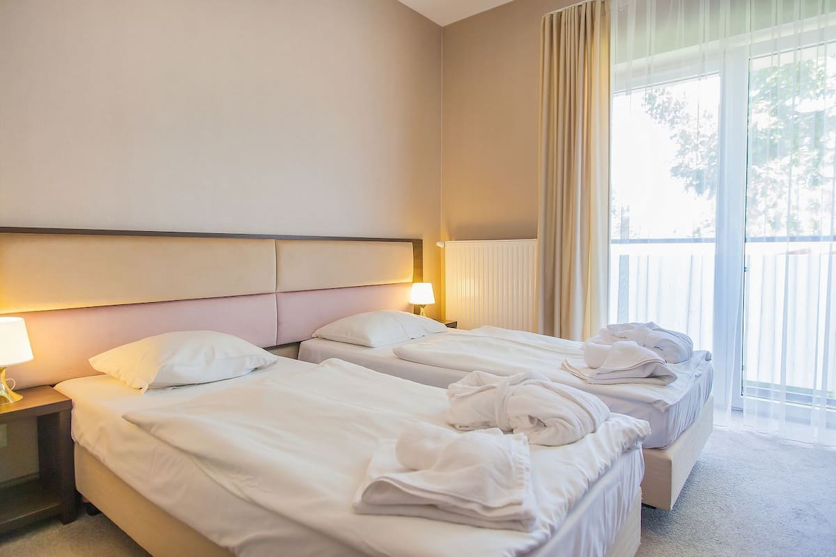 Habitación de hotel con dos camas, una mesita de noche con lámpara y vistas a un balcón.