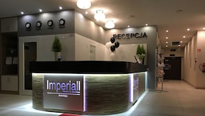 Ein Empfangsschalter mit der Aufschrift 'Imperial Resort & MediSpa', Uhren, die verschiedene Städte anzeigen, ein Empfangsbereich und ein roter Teppich.
