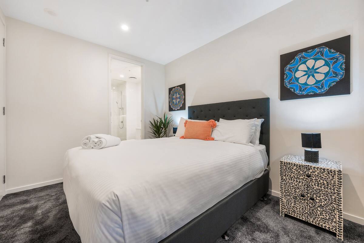 Un lit bien fait, avec du linge de lit blanc et un oreiller orange, une tête de lit sombre et une table de chevet à motifs, sur laquelle se trouve une lampe.