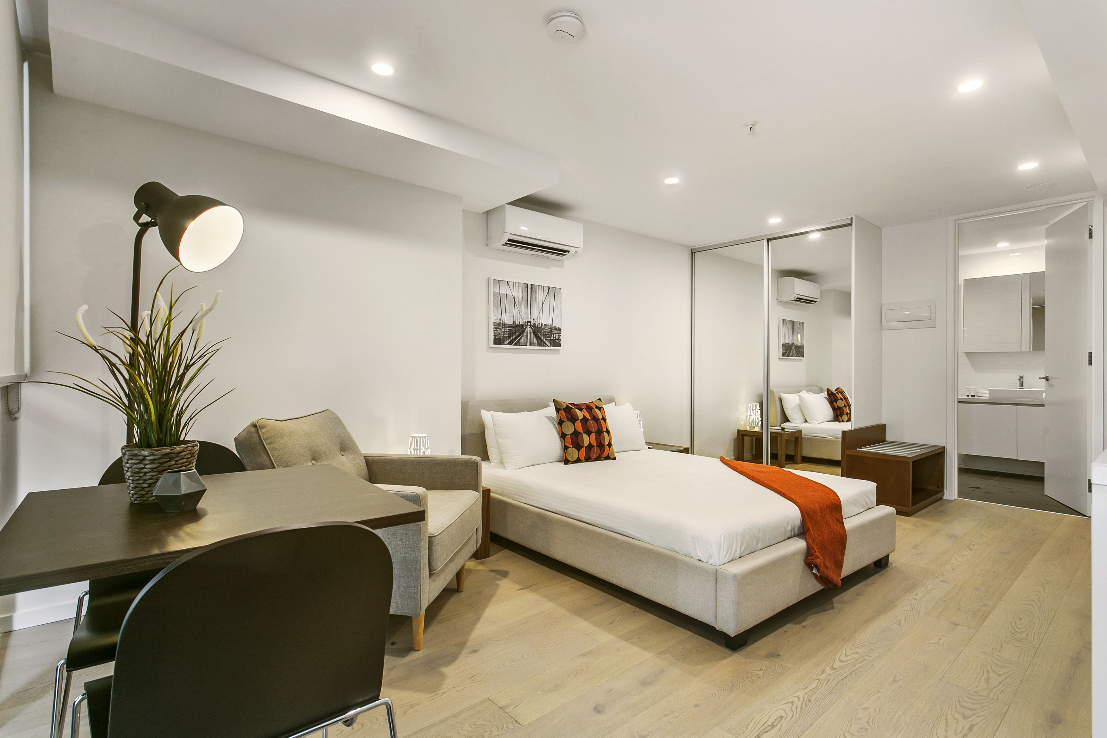 Una habitación de hotel moderna con cama, mesita de noche, lámpara, silla y un pequeño escritorio con una planta.