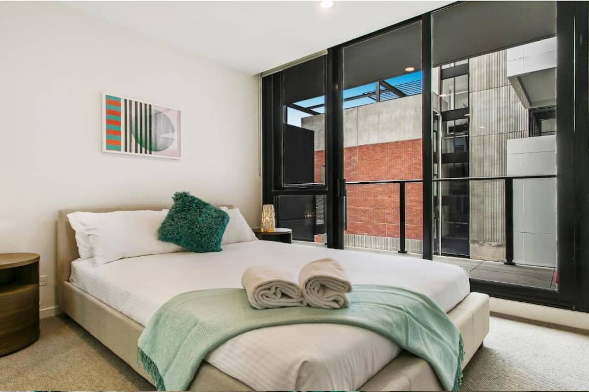 Une chambre d’hôtel moderne avec un grand lit, une vue sur la ville grâce à de grandes fenêtres du sol au plafond, une petite table d’appoint et une œuvre d’art accrochée au mur.