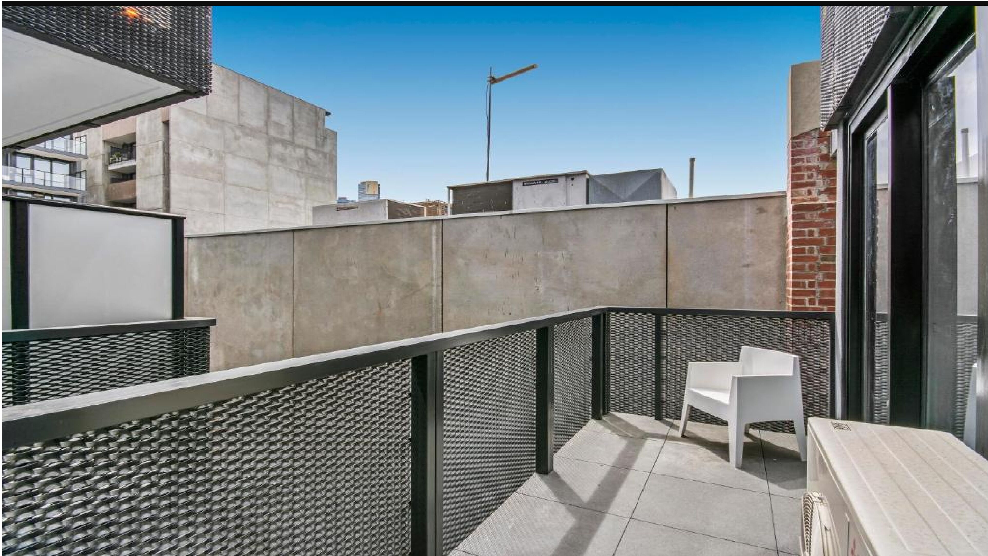 A modern outdoor balcony with a metal mesh railing, a small seating area with a chair, and a concrete wall in the background.