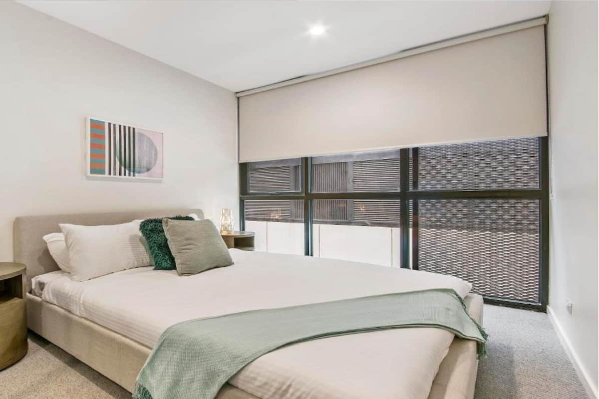 Une chambre moderne avec un grand lit, une table de chevet, une œuvre d’art accrochée au mur et une fenêtre avec des stores.