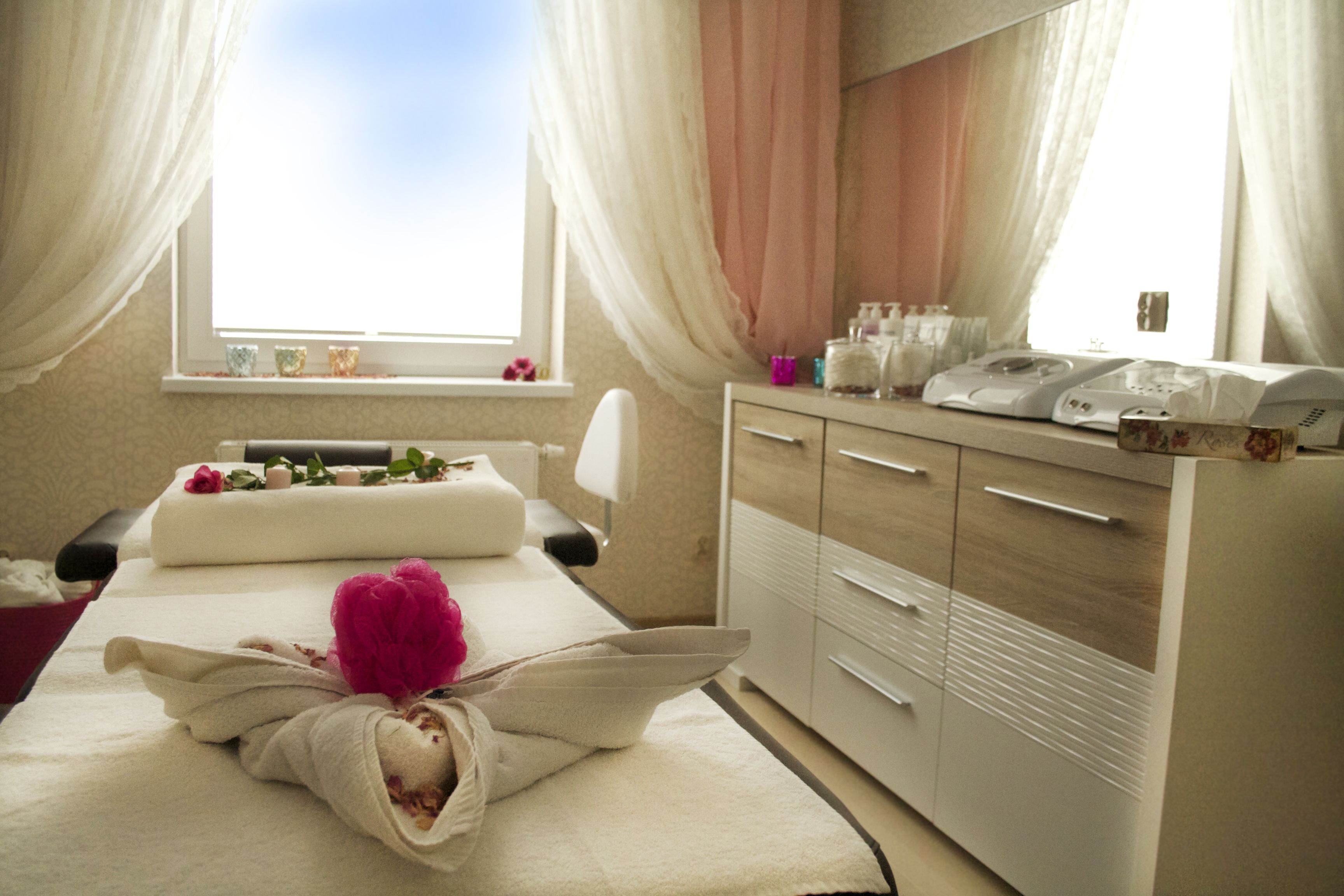 A spa room with a massage table, a massage chair, a cabinet with toiletries, and a window with curtains.