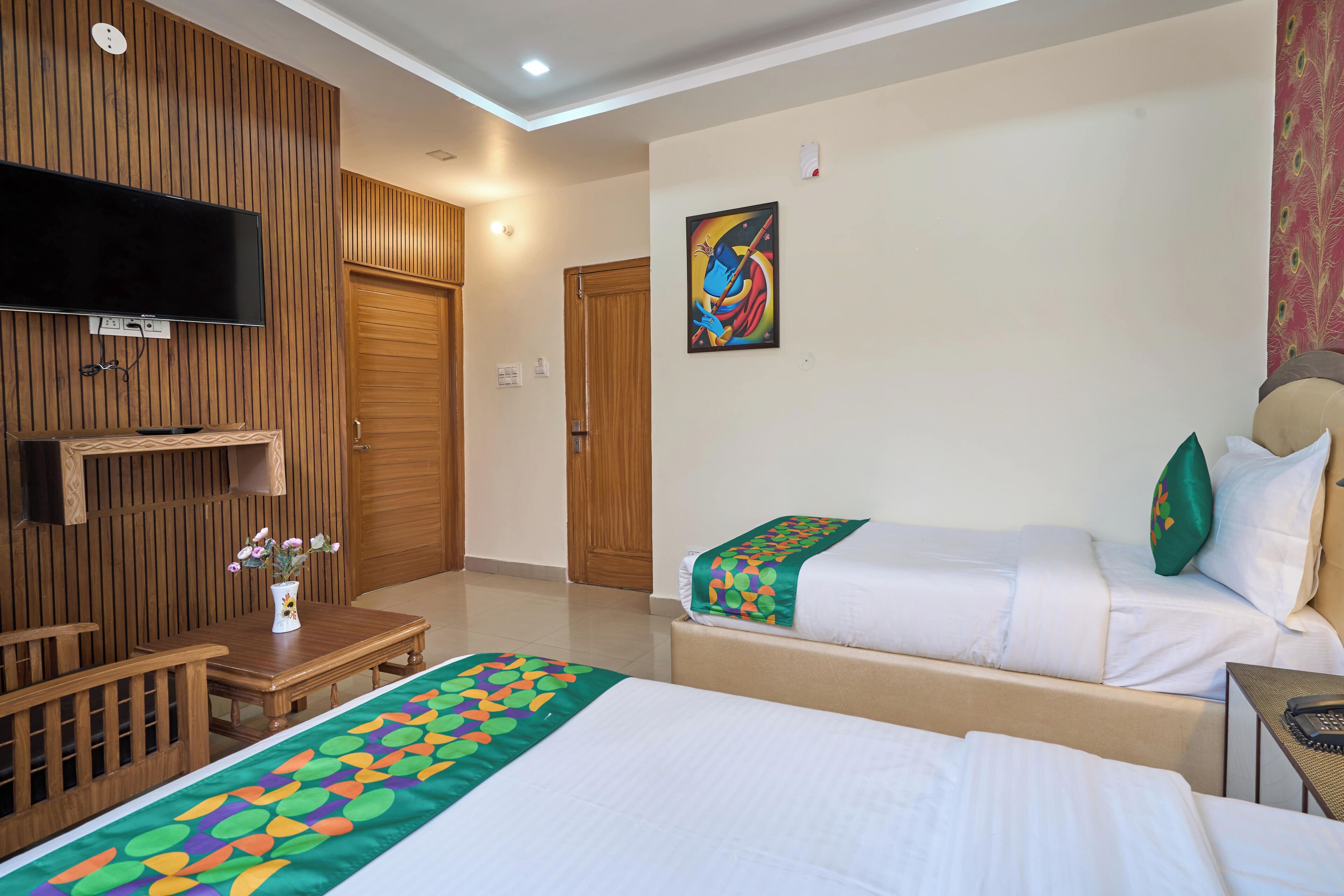 A hotel room with two beds, a TV, a wooden panel wall, a painting, and a small table with a vase.