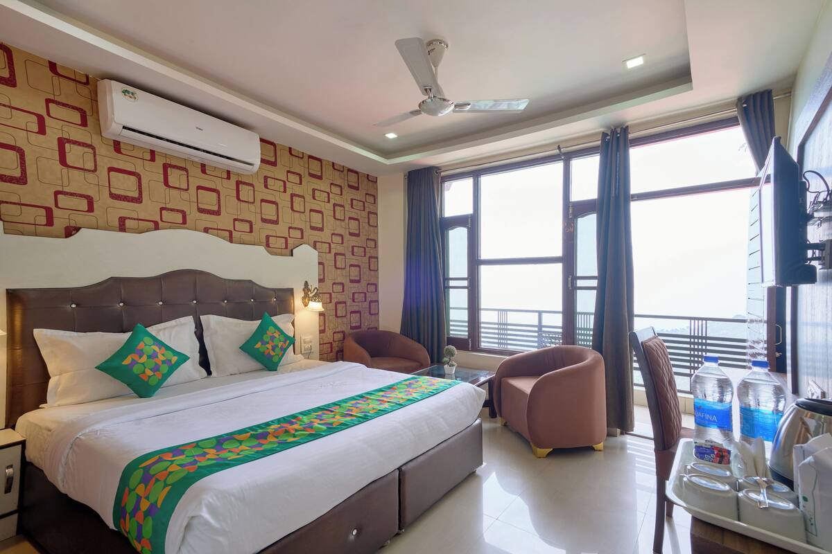 A hotel room with a bed, a ceiling fan, an air conditioner, a window with curtains, a TV, and a table with water bottles and glasses.