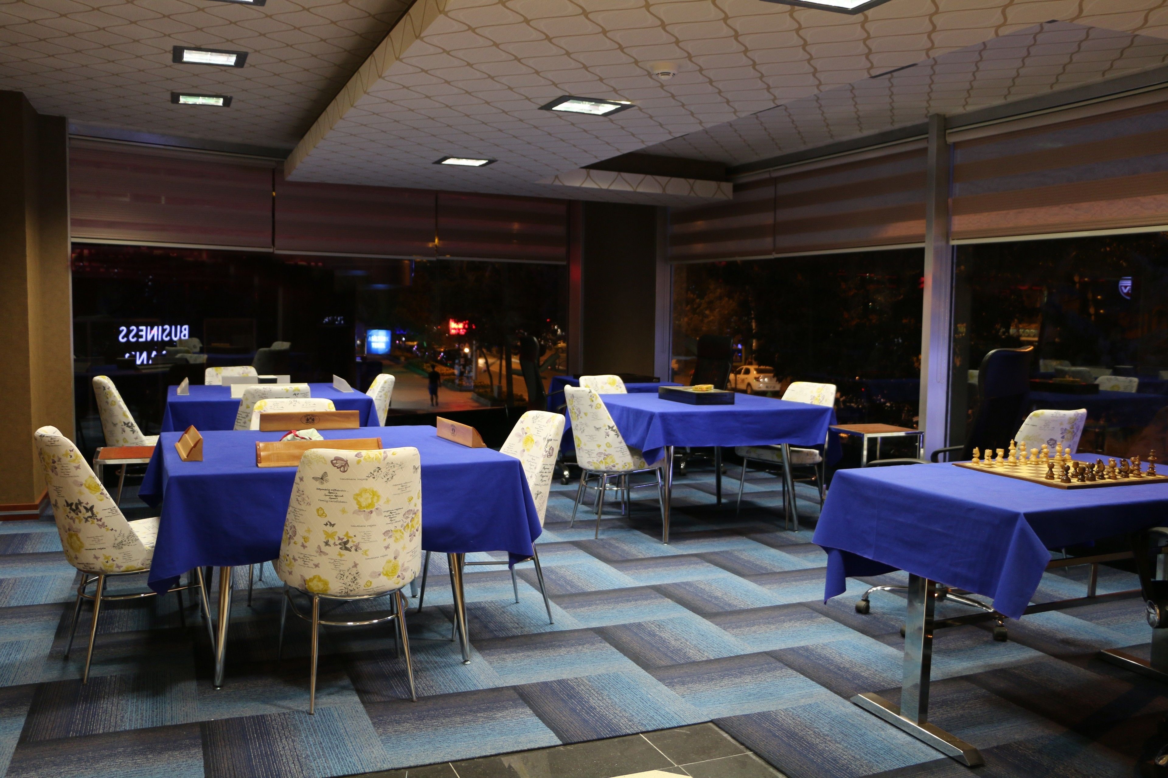 Un espace repas avec des tables recouvertes de nappes bleues, des chaises à motifs floraux et un jeu d’échecs posé sur l’une des tables.