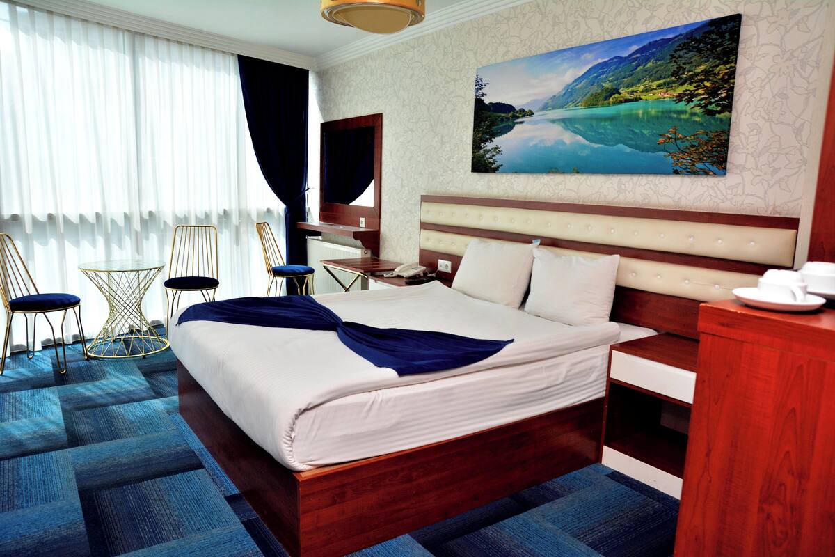 Quarto de hotel com uma cama grande, uma escrivaninha, uma cadeira, uma mesa pequena e um quadro na parede.