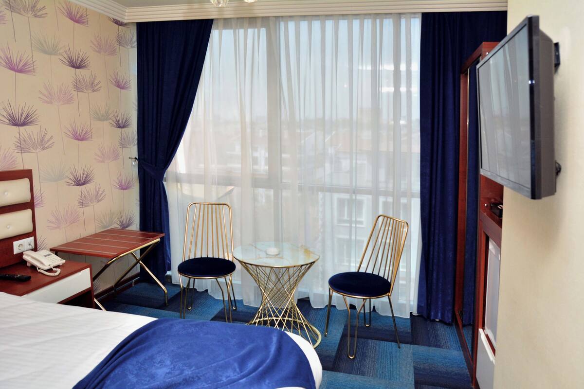 Quarto de hotel com uma cama, uma mesa com telefone, duas cadeiras, uma mesa pequena e uma televisão.