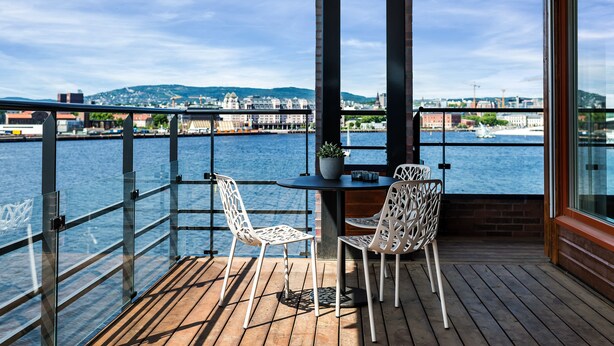 A balcony with a wooden floor, two white chairs, a small table, and a potted plant. - Wright Apartments (Oslo)