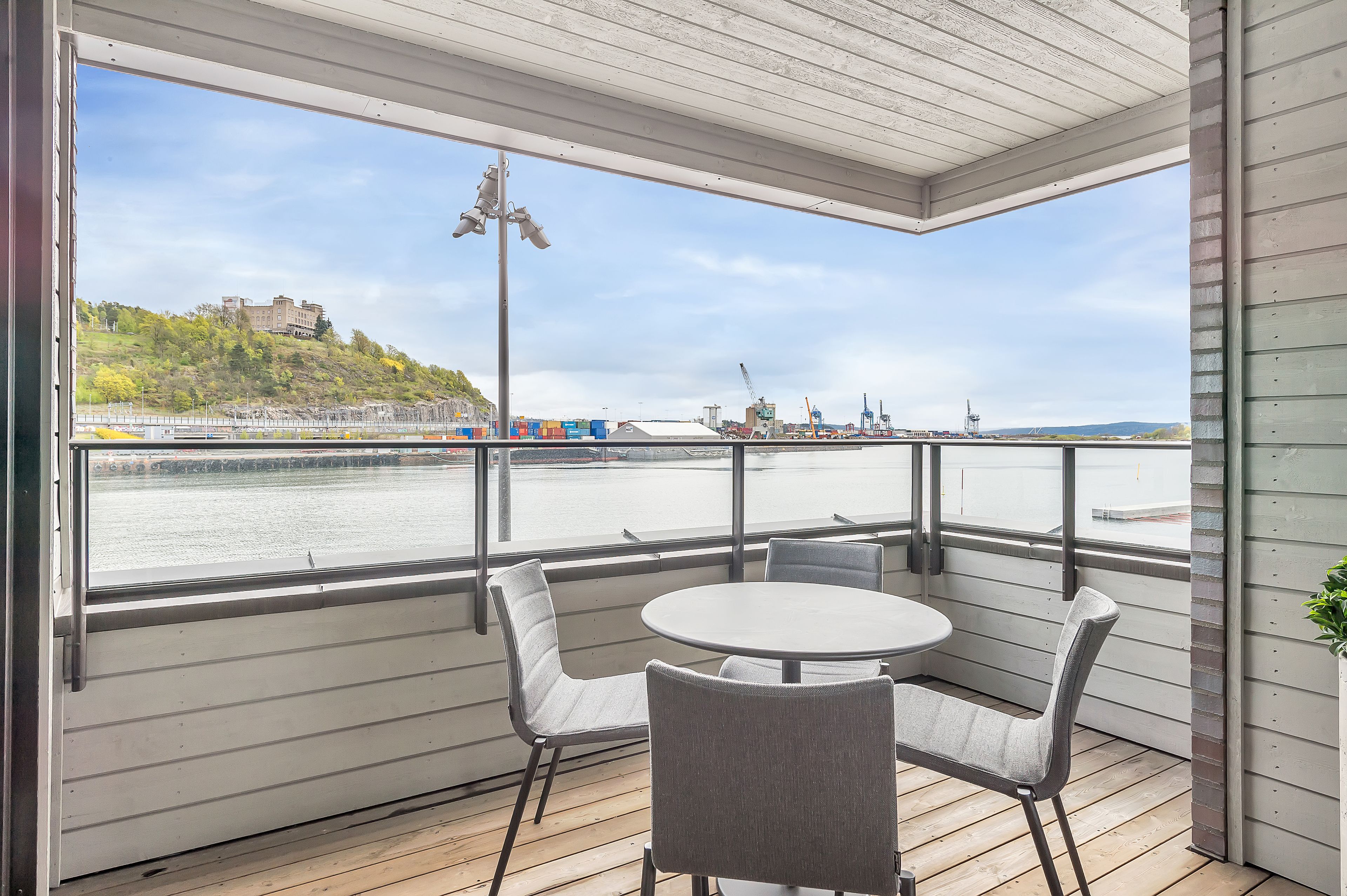 Een overdekt terras met een tafel en stoelen, met uitzicht op een waterkant, een gebouw en industriële gebouwen in de verte.