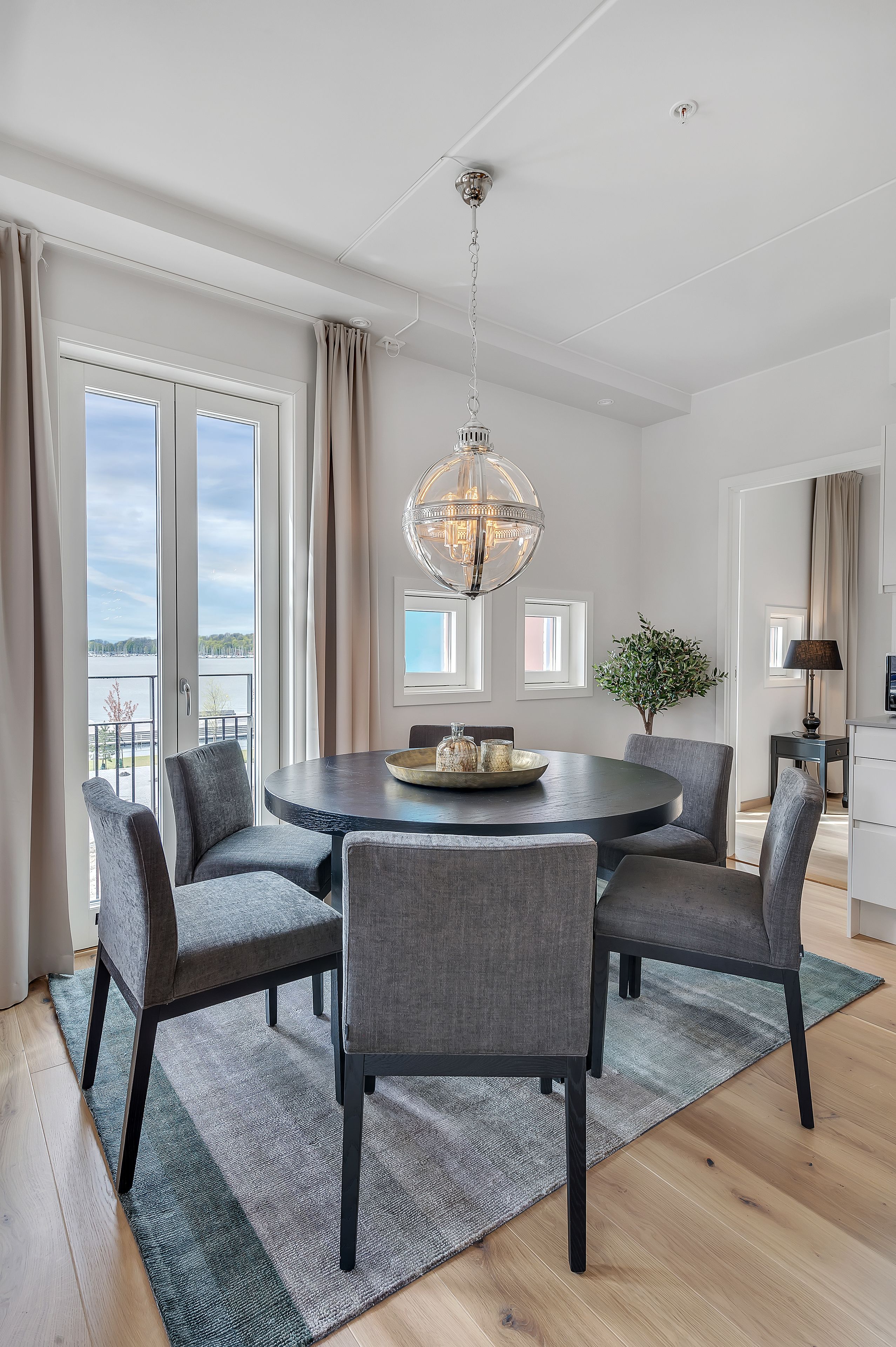 A modern dining area with a round table, four grey chairs, a chandelier, and a view of a lake through large windows.