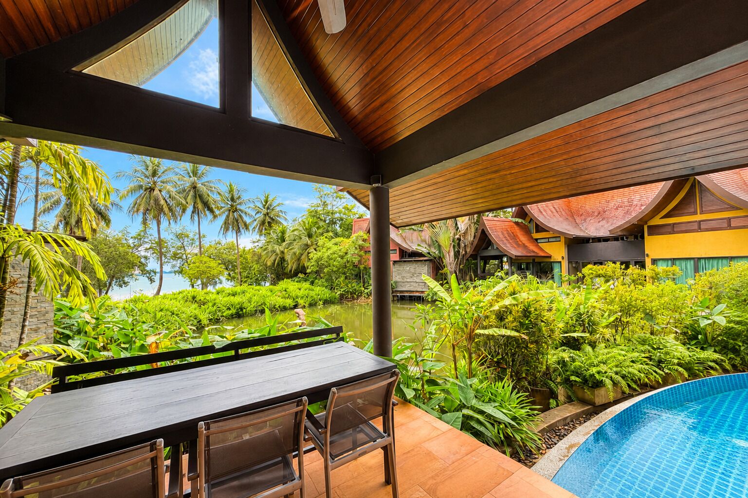 Un espacio al aire libre cubierto con piscina, mobiliario de madera y vegetación tropical.