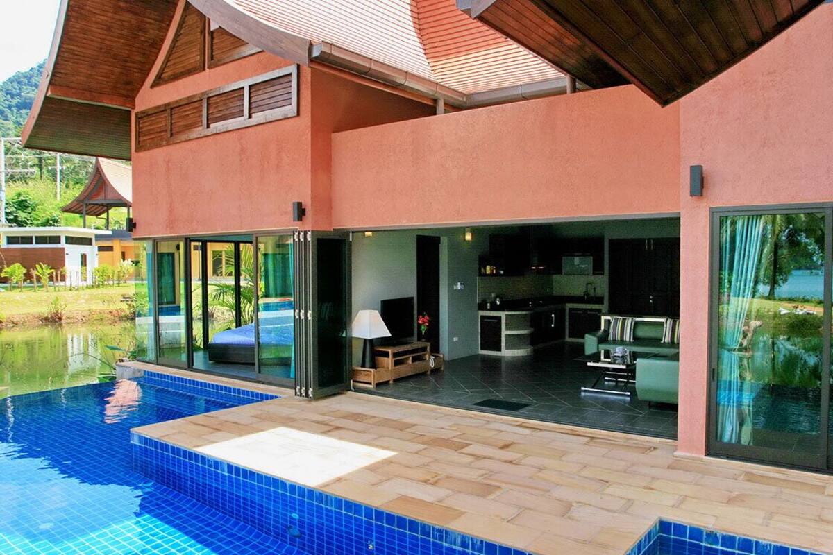 Ein modernes Haus mit Swimmingpool, großen Glasfenstern, die in einen Wohnbereich führen, und einer durch die Fenster einsehbaren Küche.
