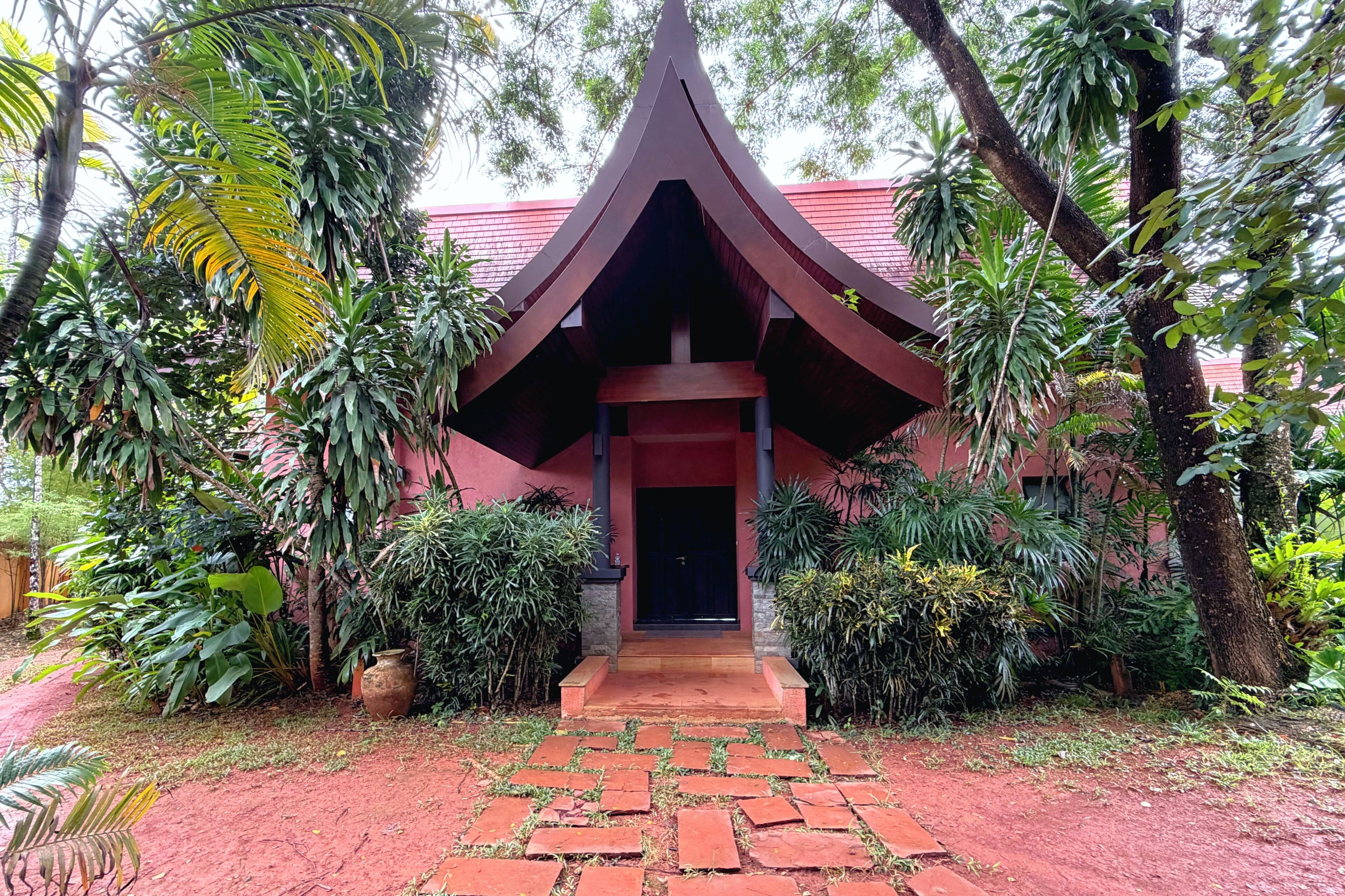 Un bâtiment doté d’une toiture à pignon remarquable, entouré d’une végétation luxuriante et accessible par un chemin pavé menant à l’entrée.