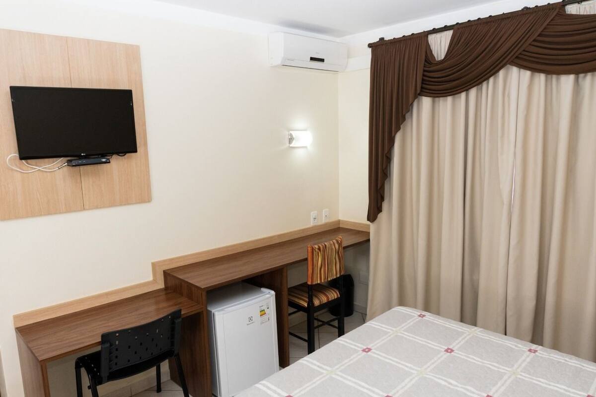 Habitación de hotel con escritorio de madera, televisor montado en la pared, refrigerador, silla y cama con colcha a cuadros.