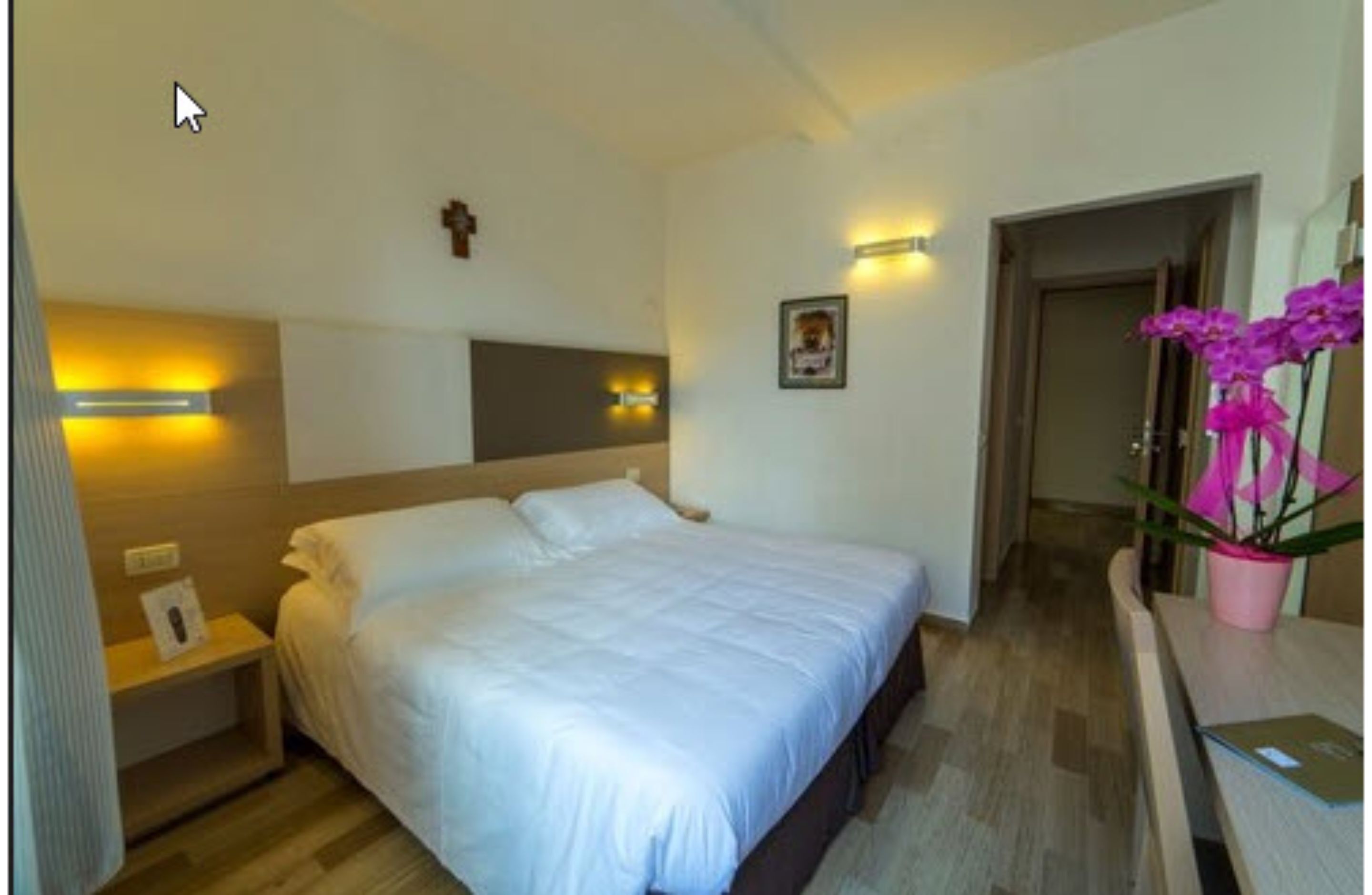 Habitación de hotel con una cama grande, mesitas de noche, una lámpara de pared, un cuadro enmarcado y una planta en maceta.
