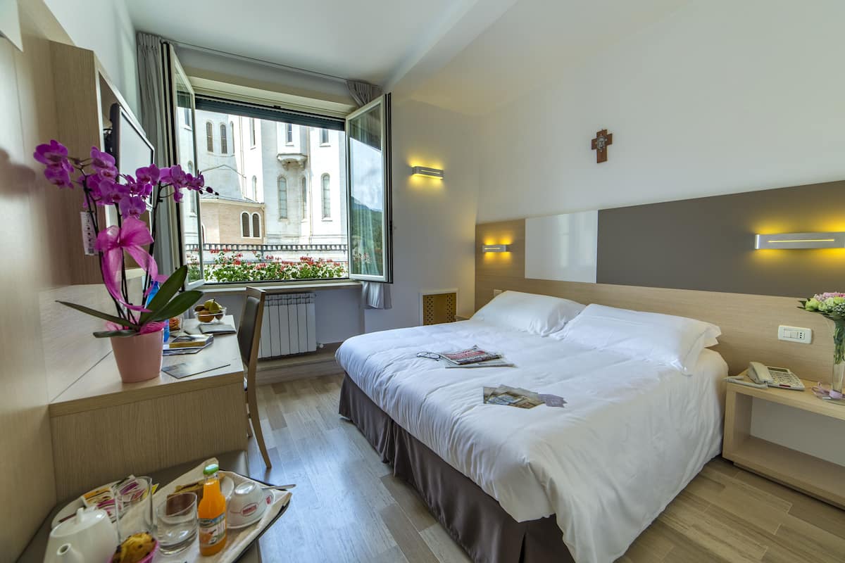 A hotel room with a large bed, a desk with a chair, a window with a view of buildings, and a tray with breakfast items.