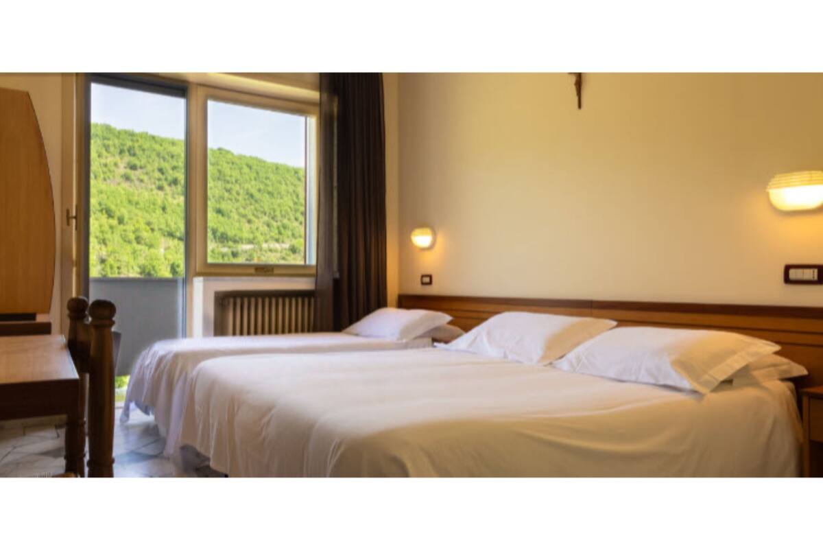 A hotel room with two beds, a wooden nightstand, a desk, and a window with a view of a green hillside.