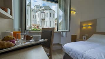Quarto de hotel com cama, mesa de jantar com itens de café da manhã, cadeira e janela com vista para um edifício.