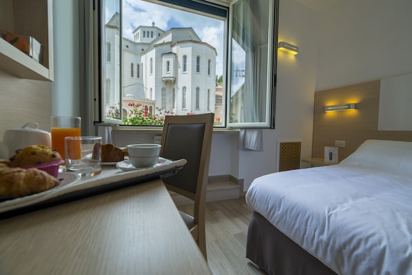 A hotel room with a bed, a dining table with breakfast items, a chair, and a window with a view of a building. - Hotel delle Rose (Cascia)