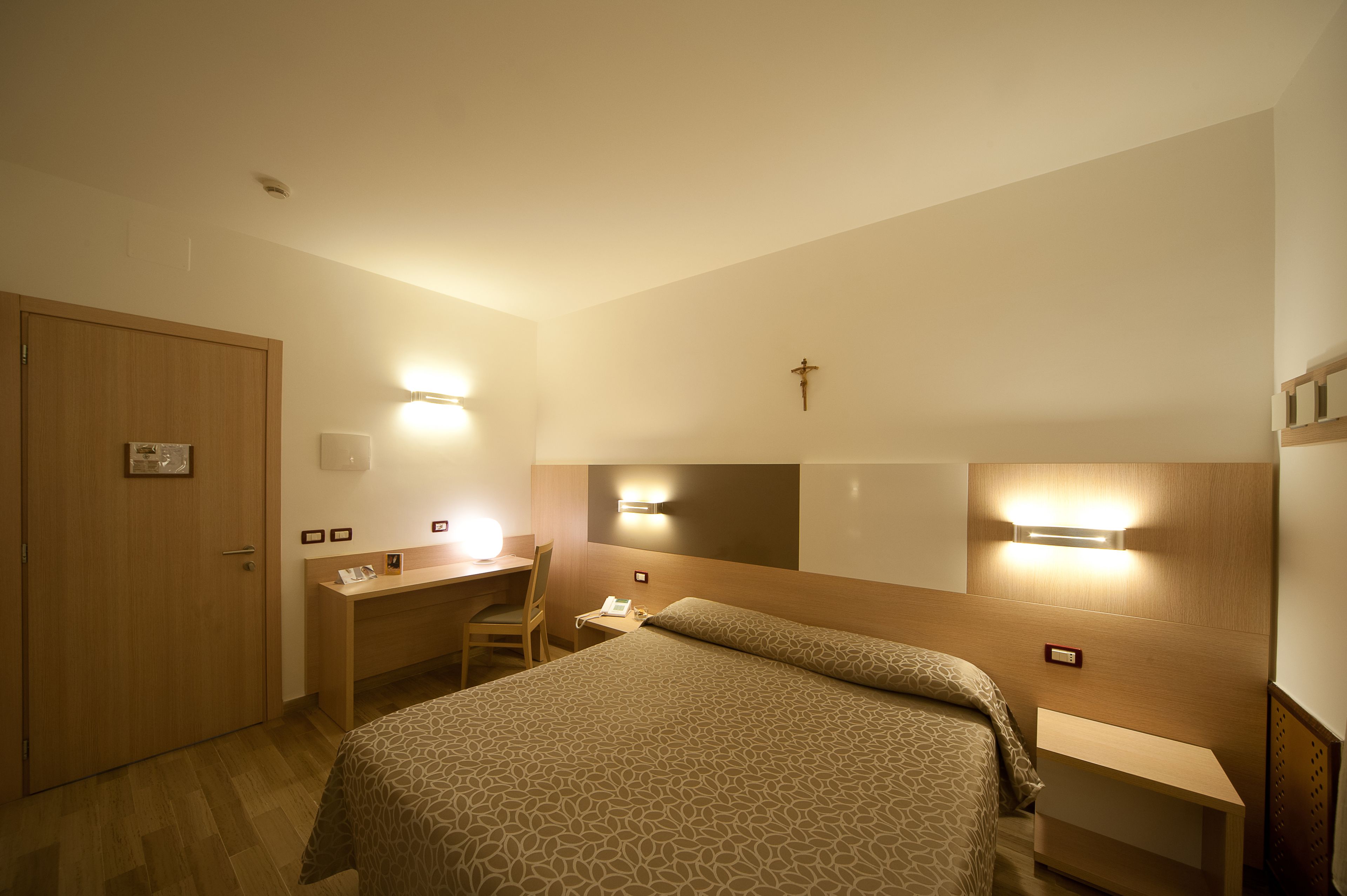 Habitación de hotel con cama, escritorio, silla y una cruz en la pared.