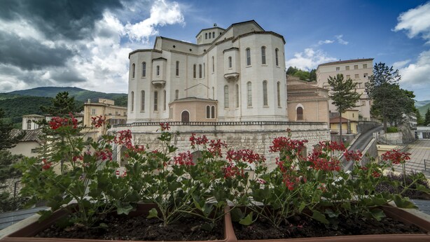 Un edificio grande e histórico, con varias secciones y un patio central, rodeado de jardines escalonados y montañas al fondo.