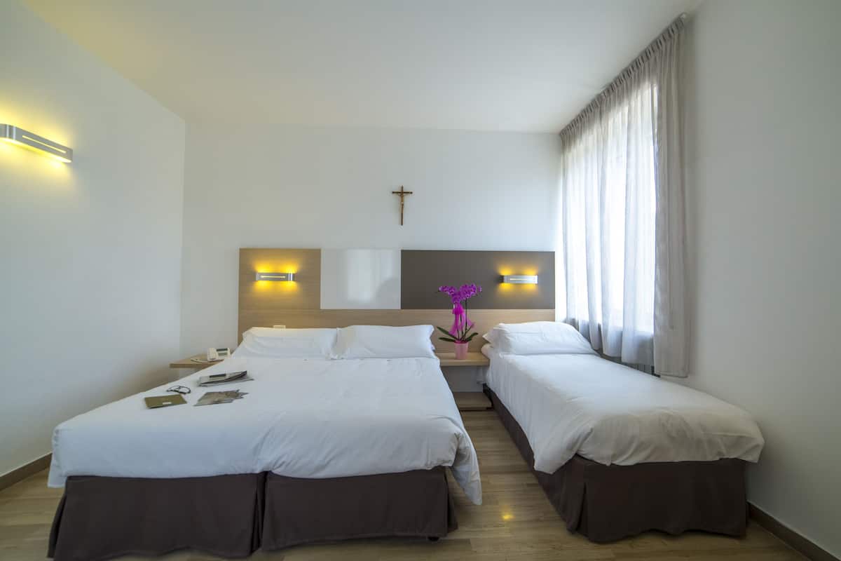 A hotel room with two beds, a nightstand, a small table with a potted plant, and a cross on the wall.