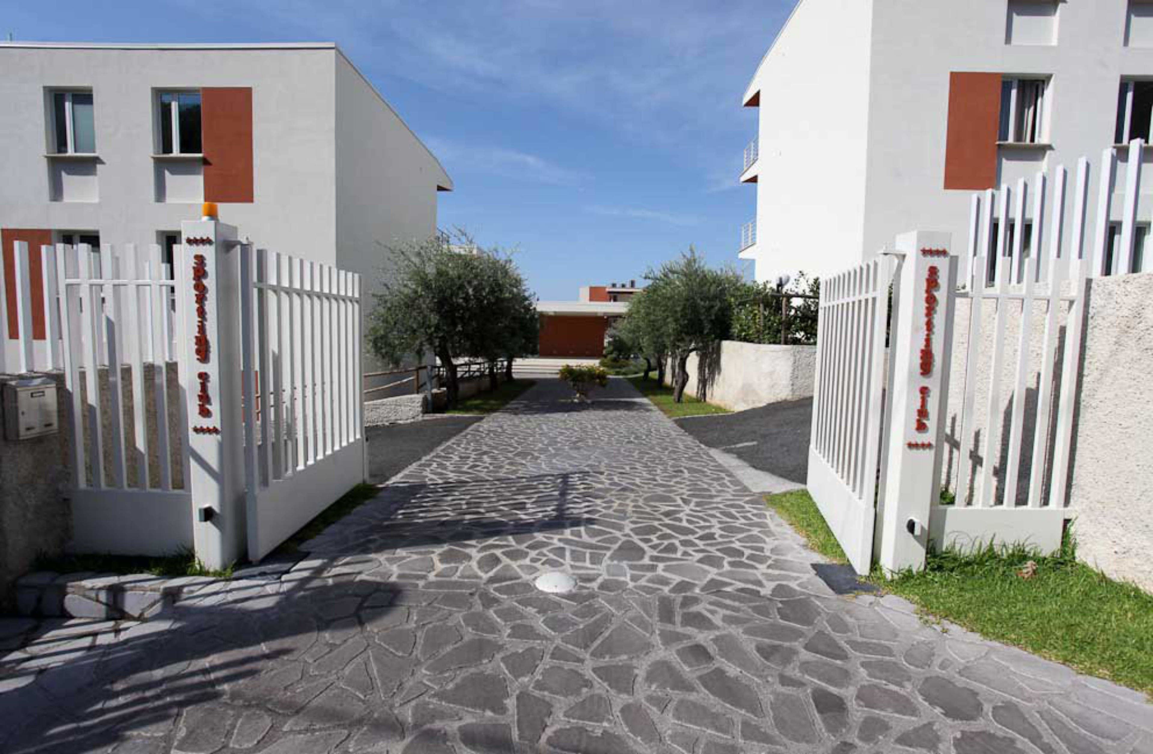 Un chemin pavé, bordé de bâtiments blancs rehaussés d’éléments rouges, mène à un bâtiment central doté d’un toit rouge.