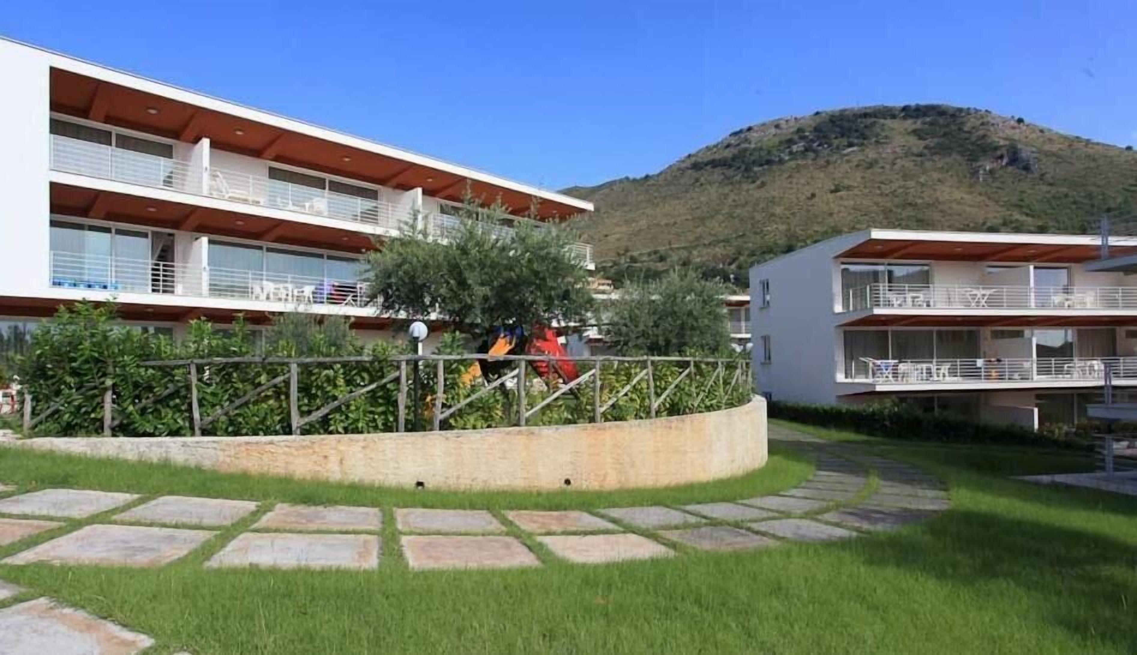 A modern two-story building with balconies, surrounded by a landscaped garden with stone pathways and greenery.