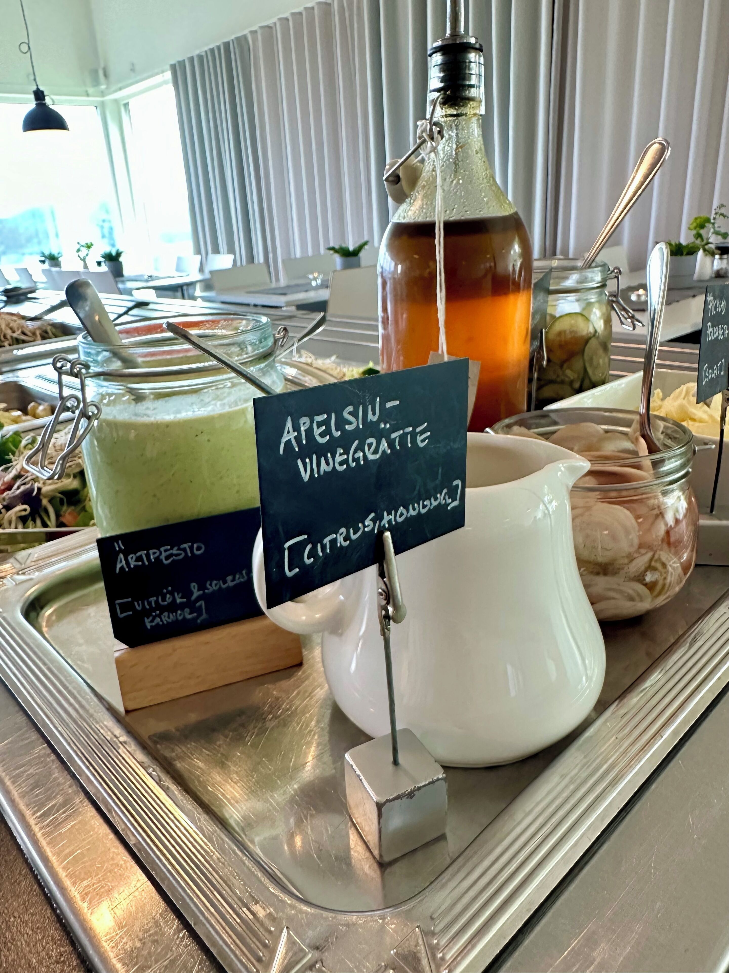 A buffet table with various food items and condiments, including a bottle of Apelsin-Vinegrätte and a pitcher of citrus juice.