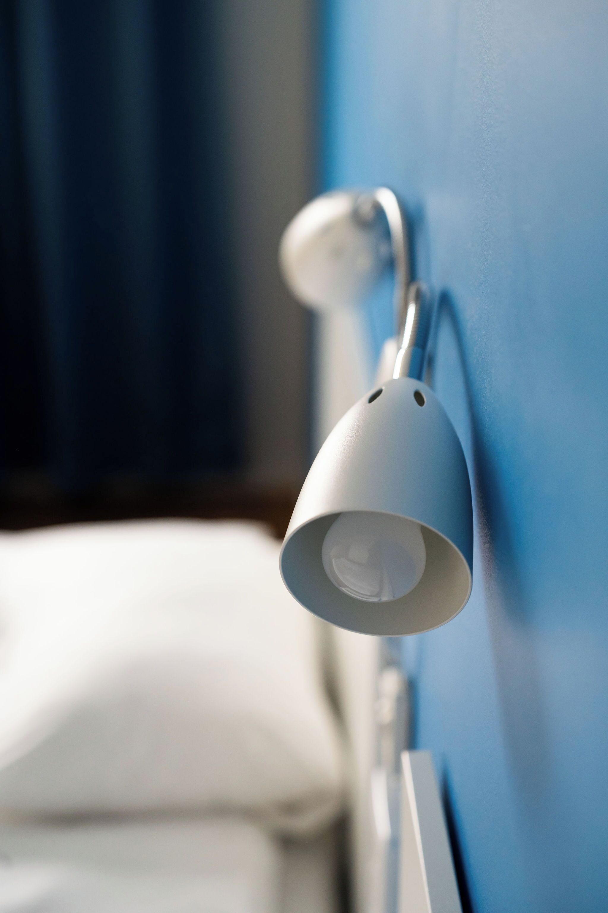 Una lámpara blanca fijada a la pared en una habitación con pared azul y cama blanca.