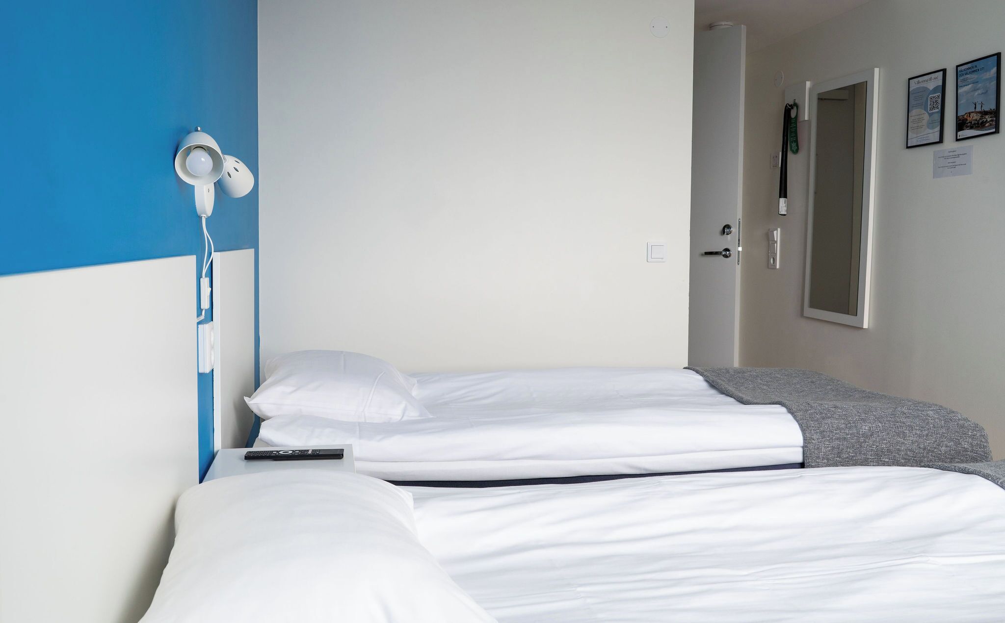 A hotel room with two beds, a blue and white wall, a bedside table with a lamp, and a door with a mirror.