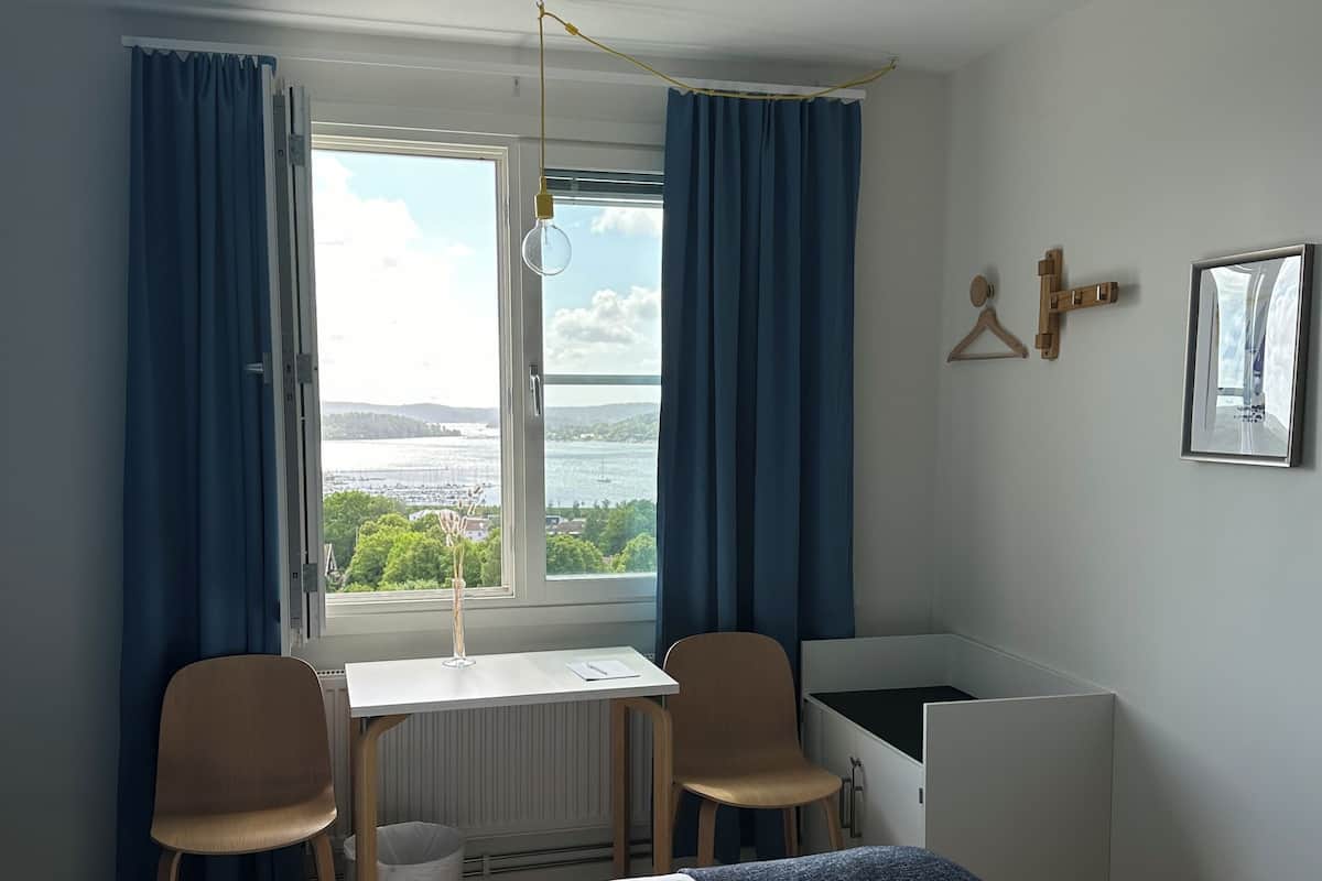 Une chambre d’hôtel comprenant un lit, un bureau, deux chaises, un radiateur, une fenêtre avec des rideaux et une vue sur la mer.
