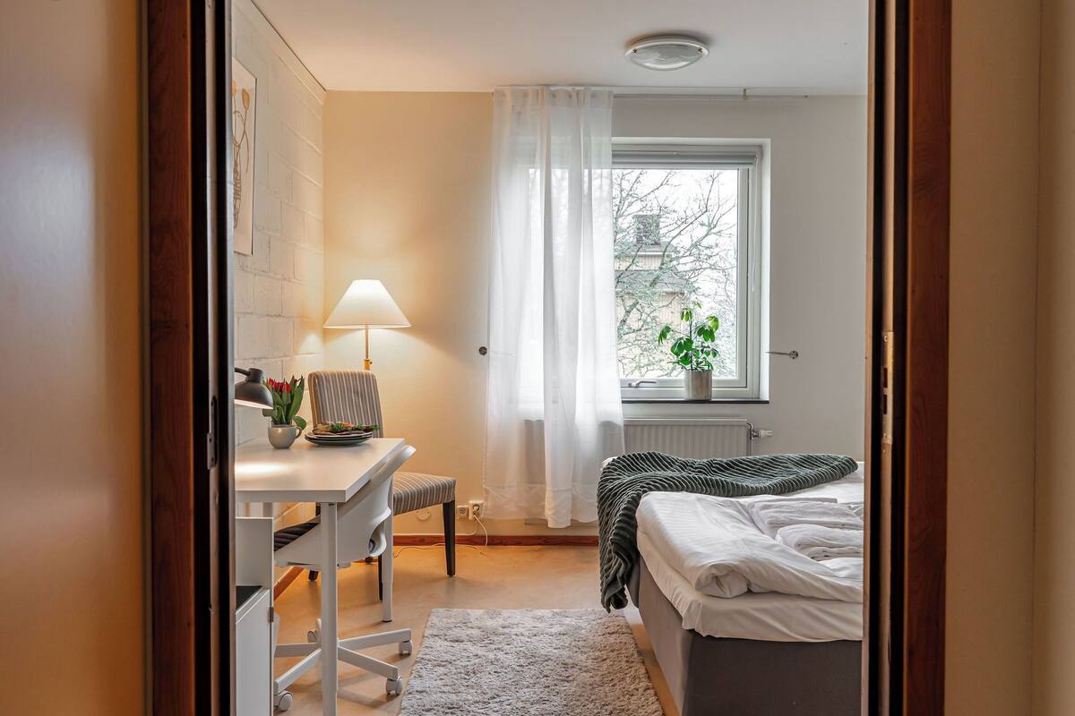 Une chambre à coucher avec un lit, un bureau avec une chaise, une lampe et une fenêtre donnant sur des arbres.