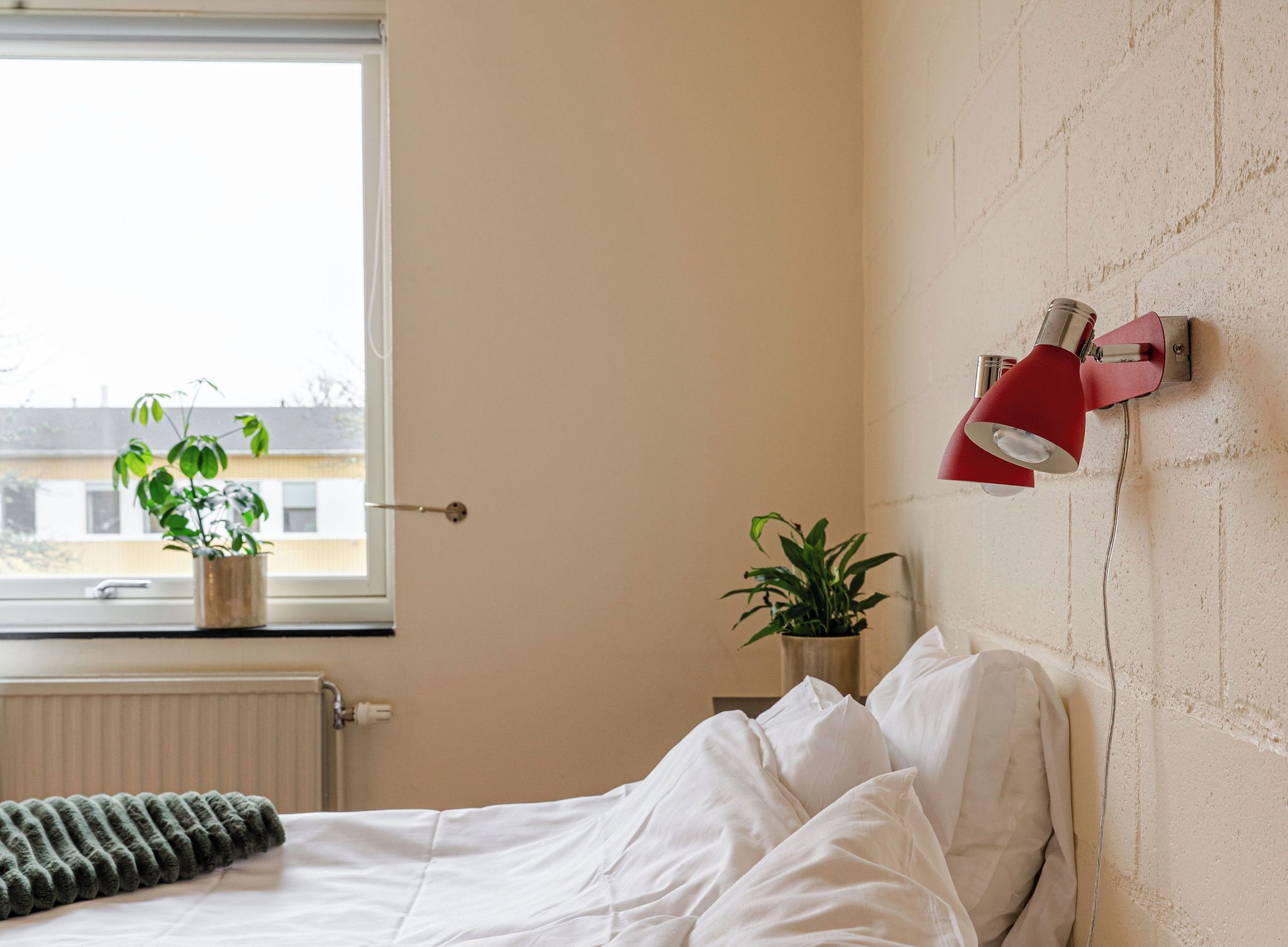 Un dormitorio con una cama, una ventana, un radiador, una planta en maceta y una lámpara de pared.