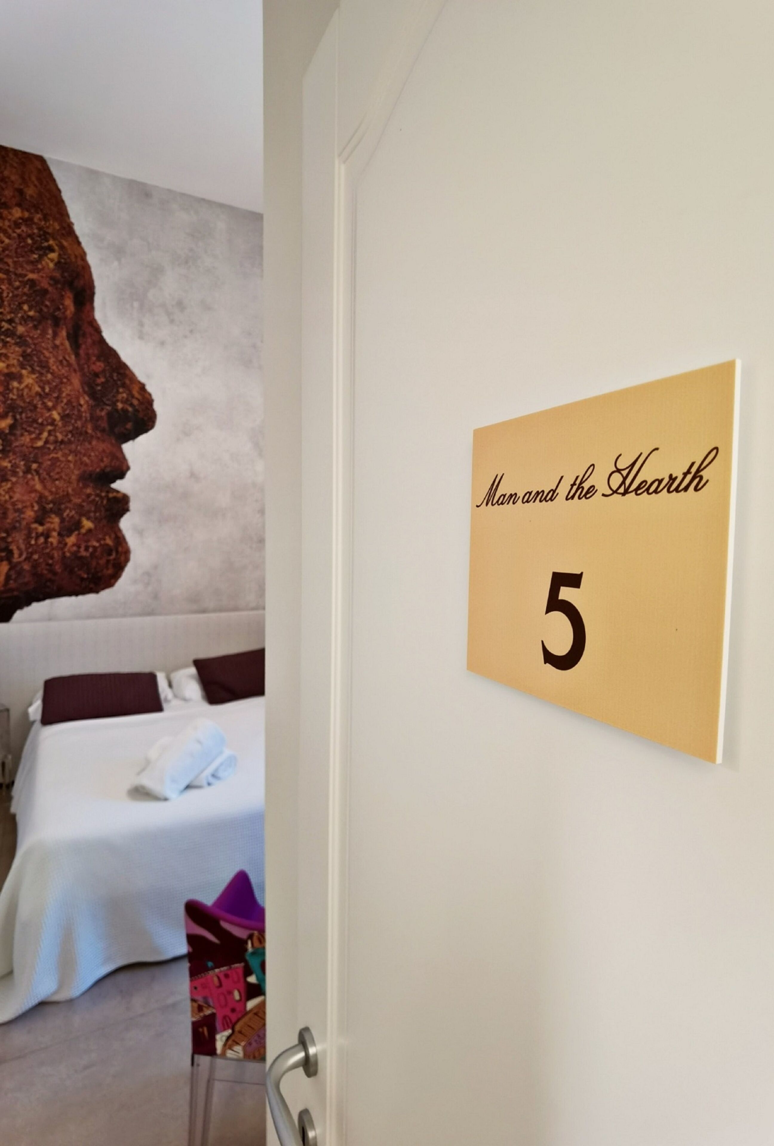 Une chambre d’hôtel avec une grande fresque murale, un lit recouvert de draps blancs et une porte sur laquelle est apposée une plaque indiquant « L’homme et le foyer 5 ».