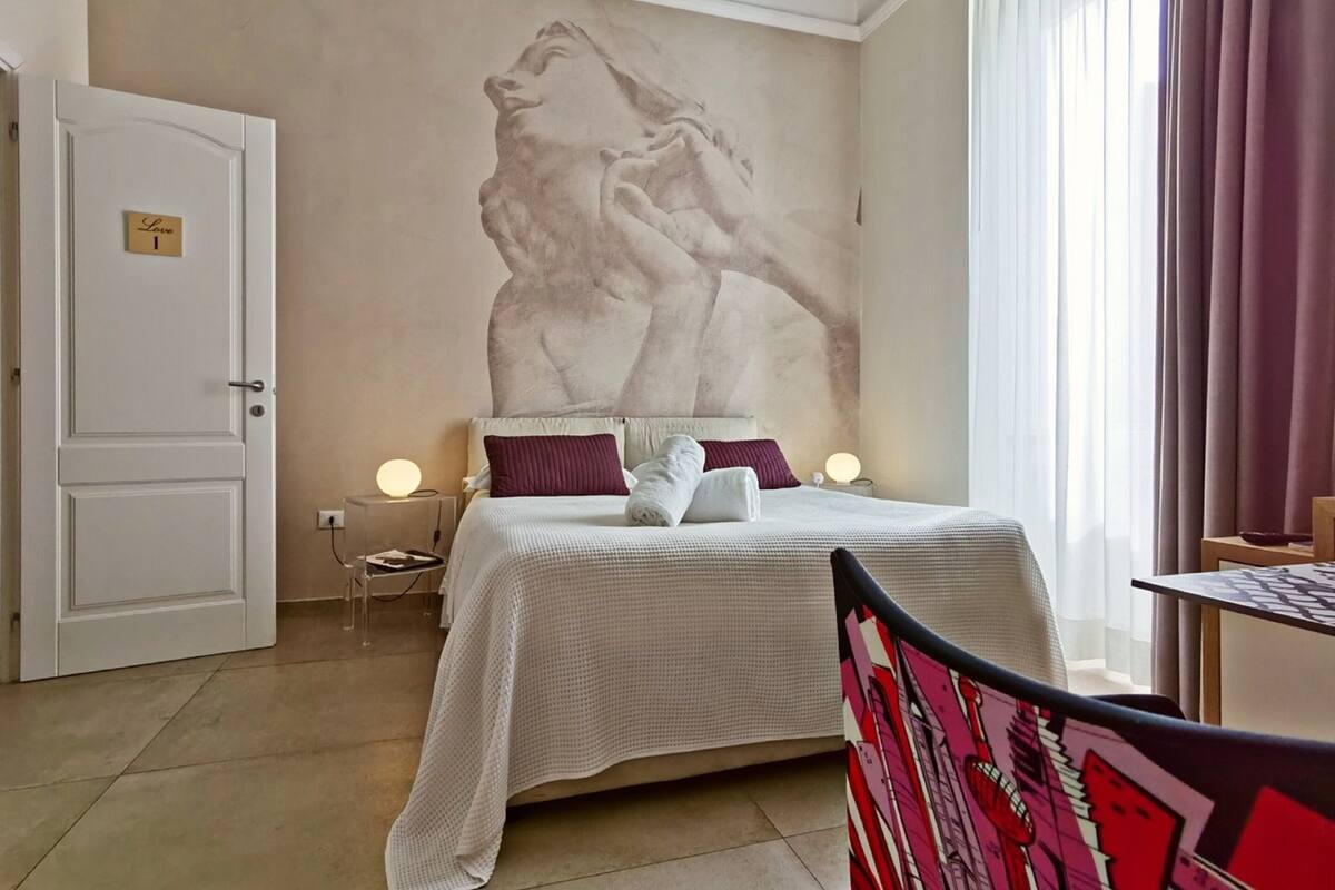 A hotel room with a bed, a wall-mounted artwork, a bedside table with a lamp, and a door labeled "Room 1".