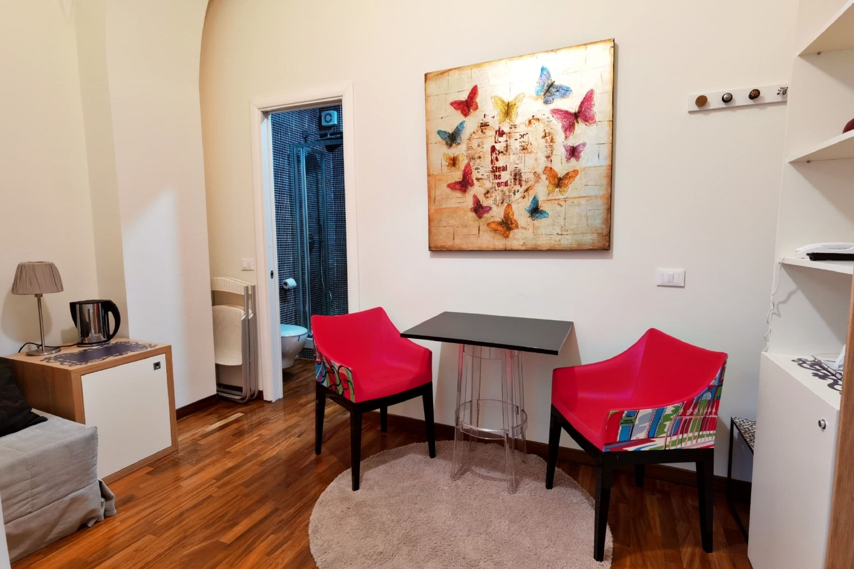 Un espacio habitable compacto con zona de comedor que incluye dos sillas rojas, una mesita y una obra de arte con forma de mariposa colgada en la pared.