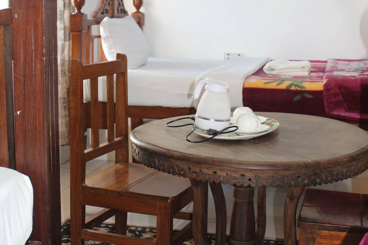 A room with a wooden bed, a wooden chair, a round table with a pitcher and cups, and a blue drape.