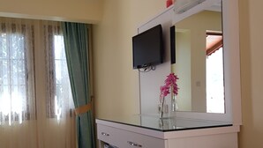 A hotel room with a white desk, a flat-screen TV, a mirror, a small vase with pink flowers, a coffee maker, and a kettle. - Ilkiz Beach Hotel (Fethiye)