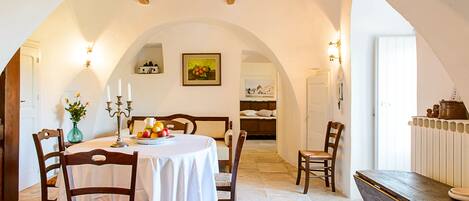 A dining area with a round table, chairs, and a white tablecloth. There is a vase with flowers on the table.