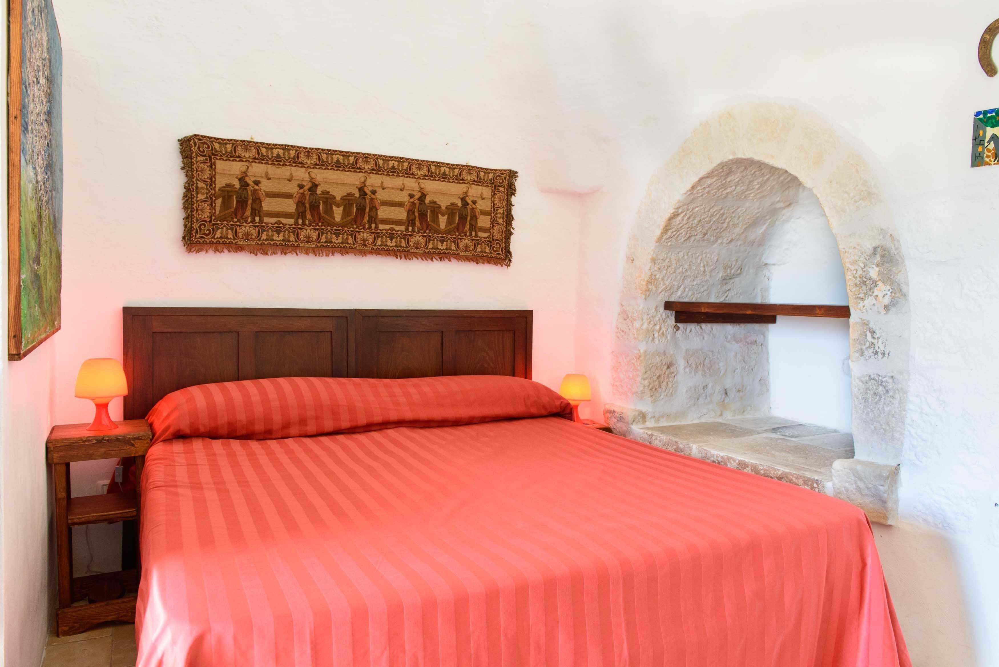 A bedroom with a wooden headboard, a red-striped bedspread, two bedside lamps, and a stone archway.
