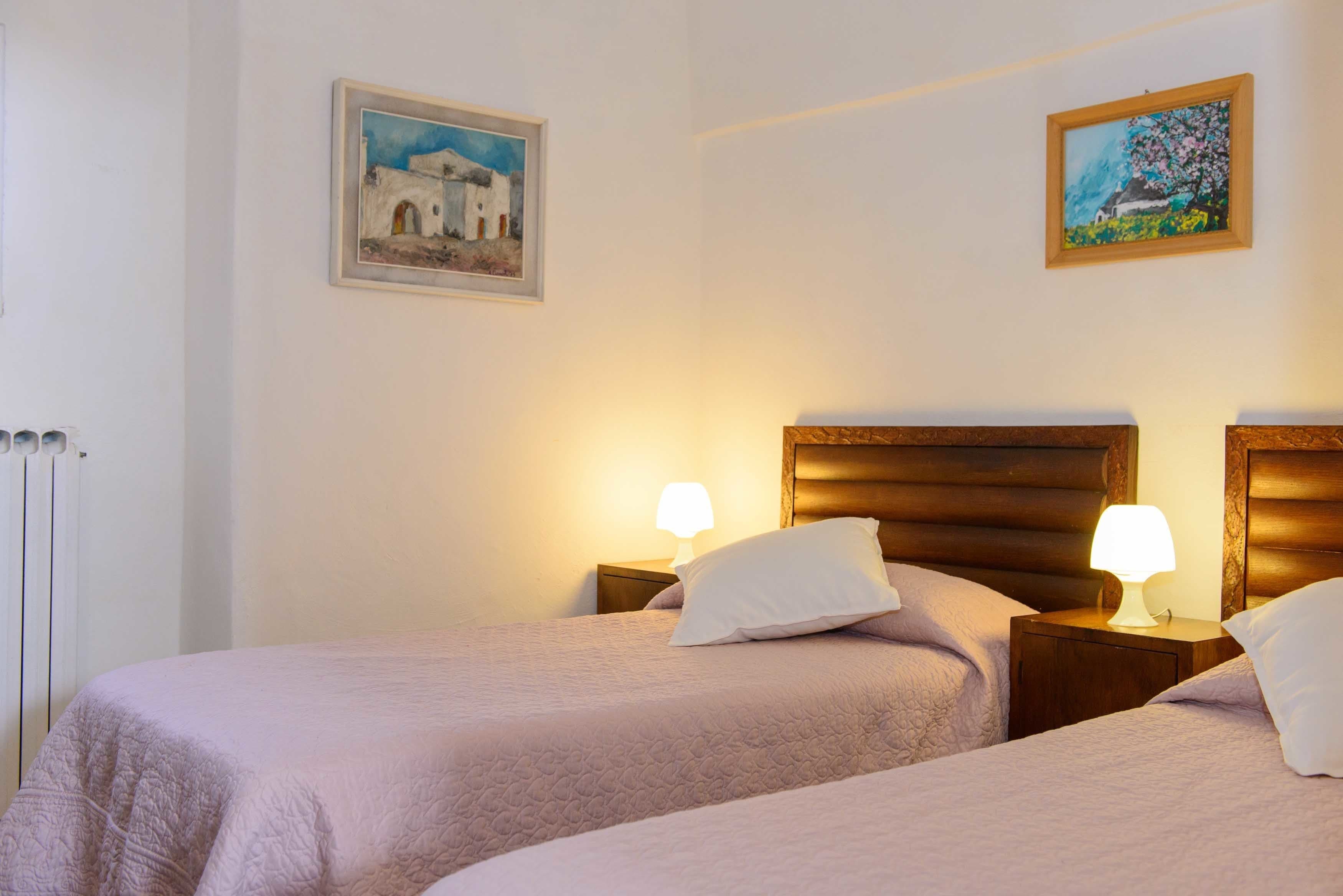 A hotel room with two beds, each with a wooden headboard and a bedside lamp. There are two framed paintings on the walls.