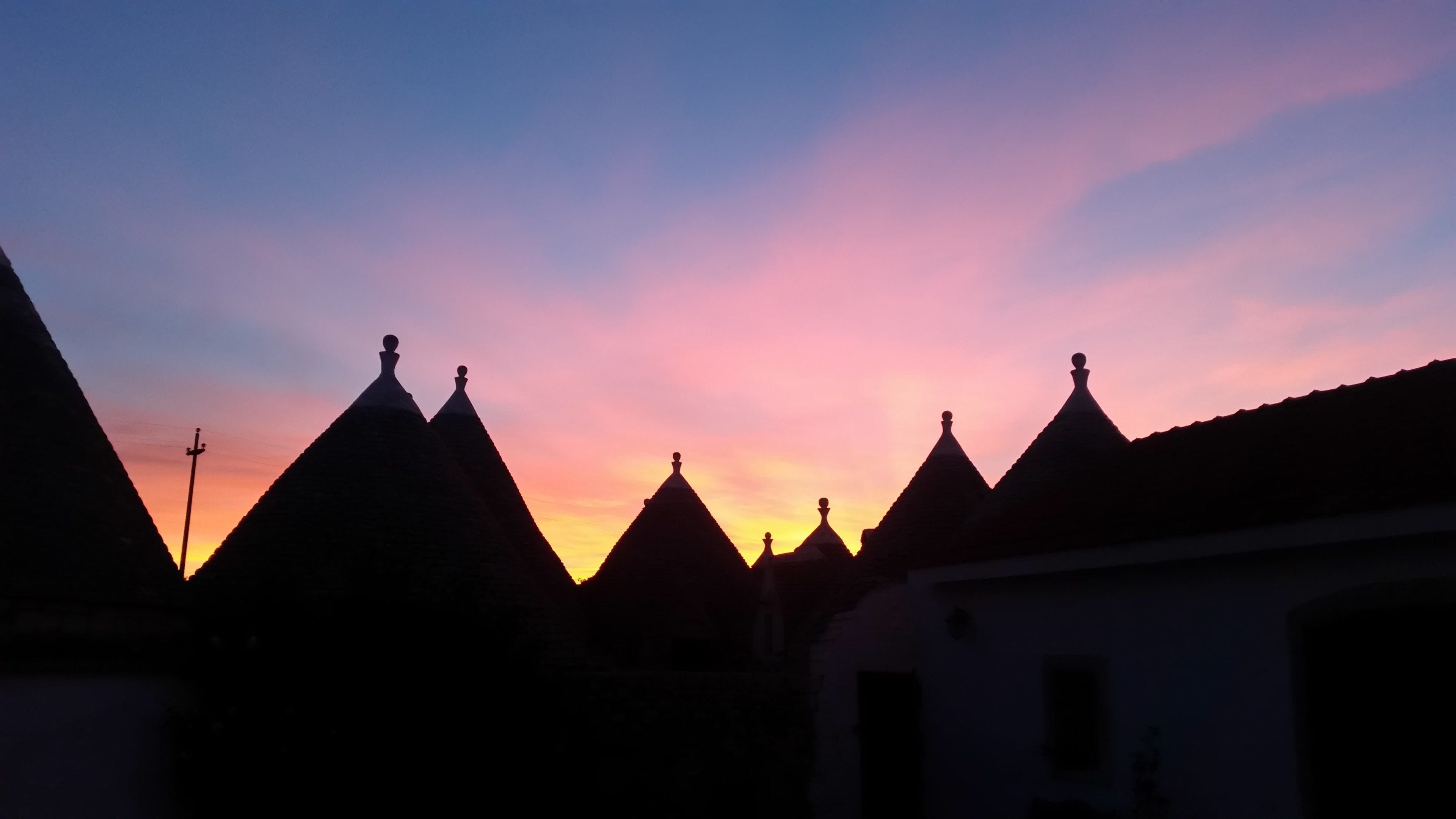 Eine Reihe von Gebäuden mit spitz zulaufenden Dächern, die vor einem Sonnenuntergangshimmel in Silhouette stehen.