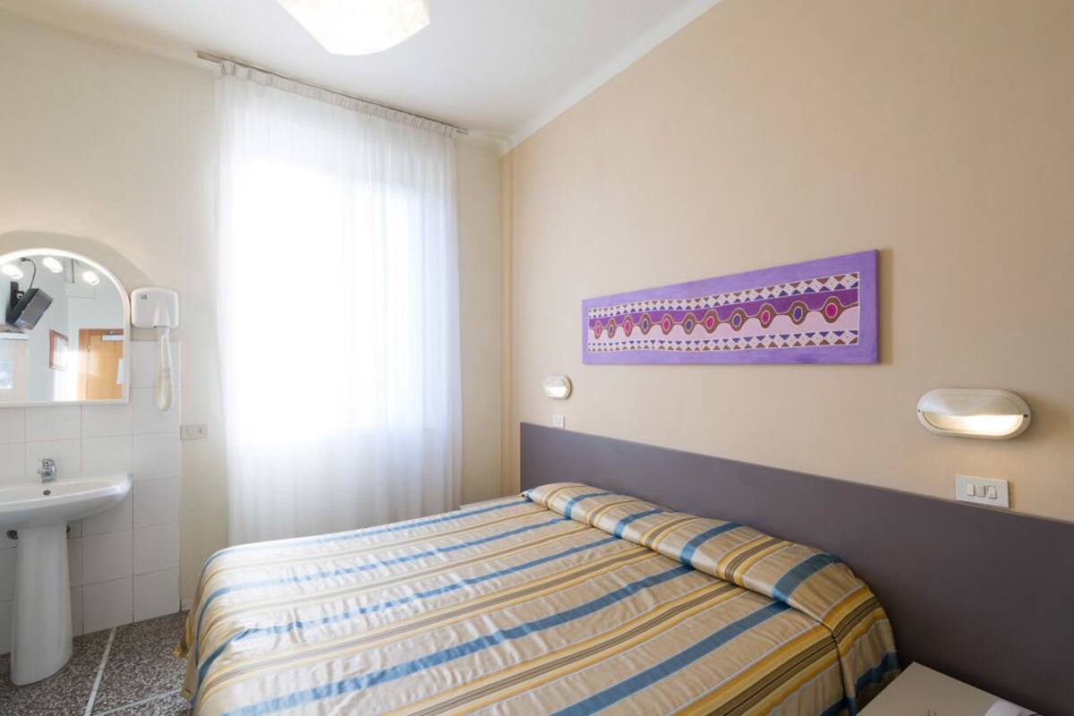 Habitación de hotel con cama, baño con lavamanos y espejo, ventana con cortinas y una lámpara de pared.