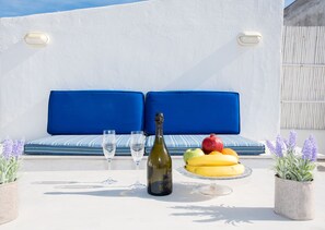 A terrace with a white wall, blue cushioned seating, a bottle of wine, glasses, and a fruit plate.