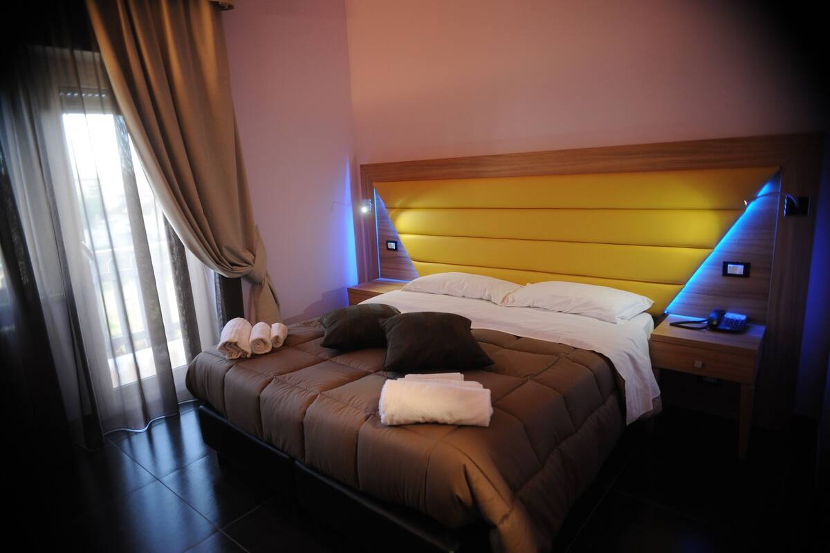 A hotel room with a large bed, a wooden headboard, a bedside table with a phone, and a window with curtains.