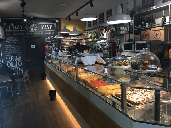 A bakery counter with various baked goods displayed in glass cases, a coffee machine, and shelves stocked with beverages and pastries.